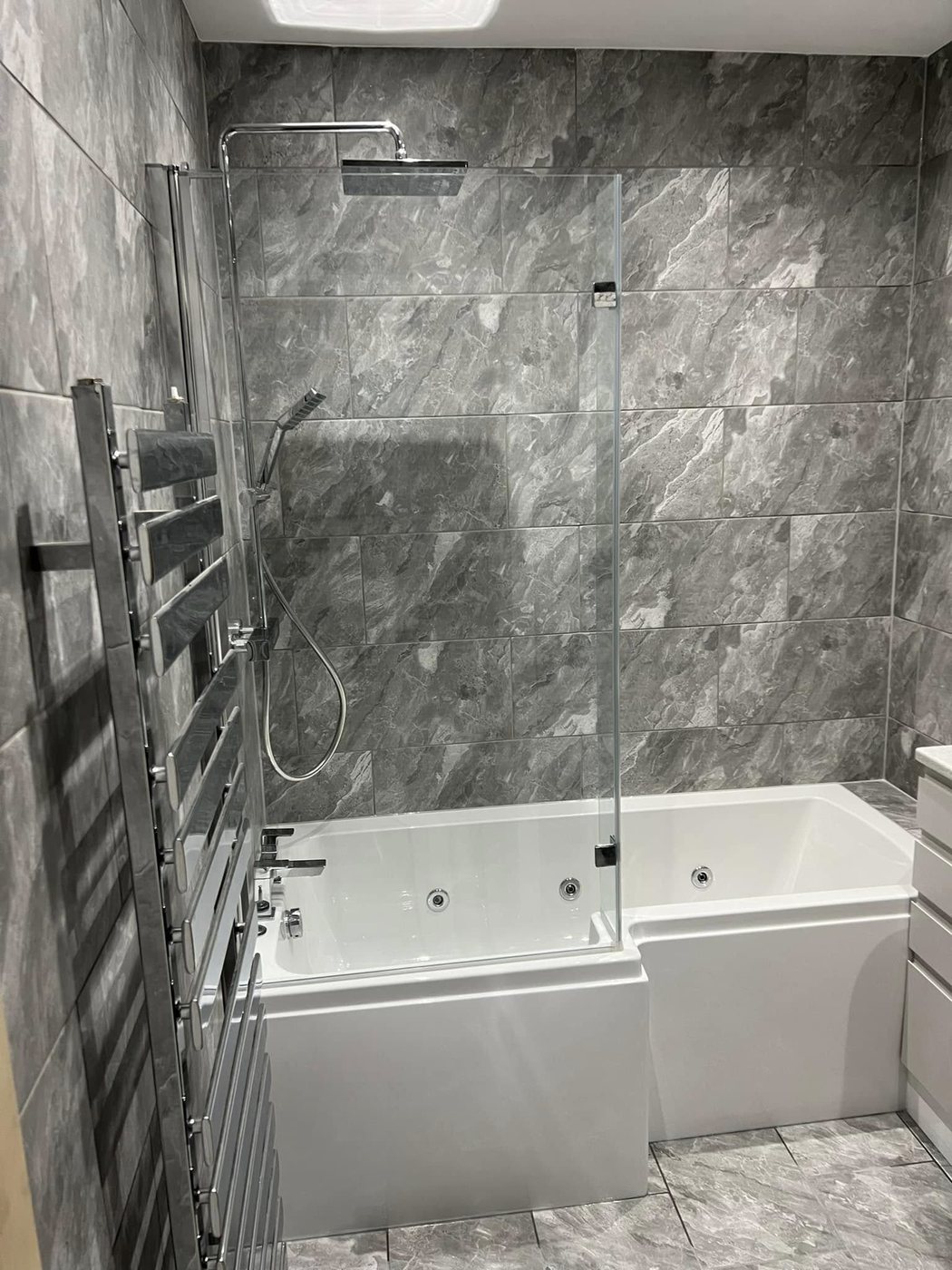 Bathroom after full NextGen renovation — grey marble-effect tiling, L-shaped bath and rainfall shower